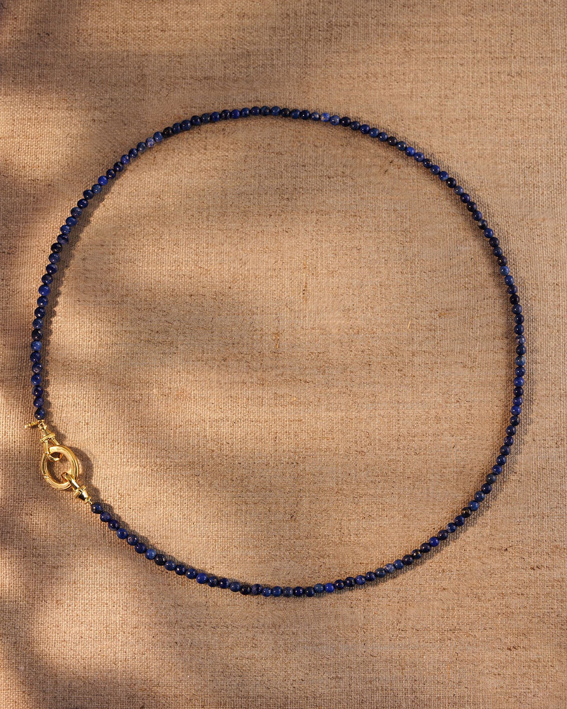 Lapis gemstone beaded necklace with gold oval clasp on neutral fabric background