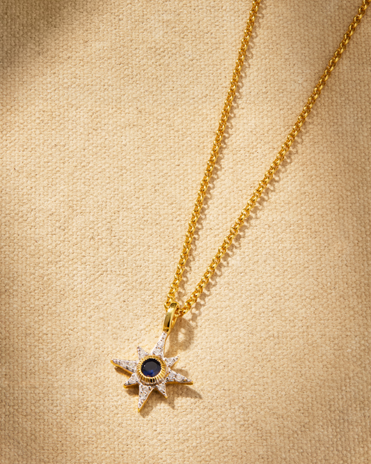A delicate NORTH STAR necklace with a sapphire blue crystal pendant on a gold chain, displayed on a textured background.
