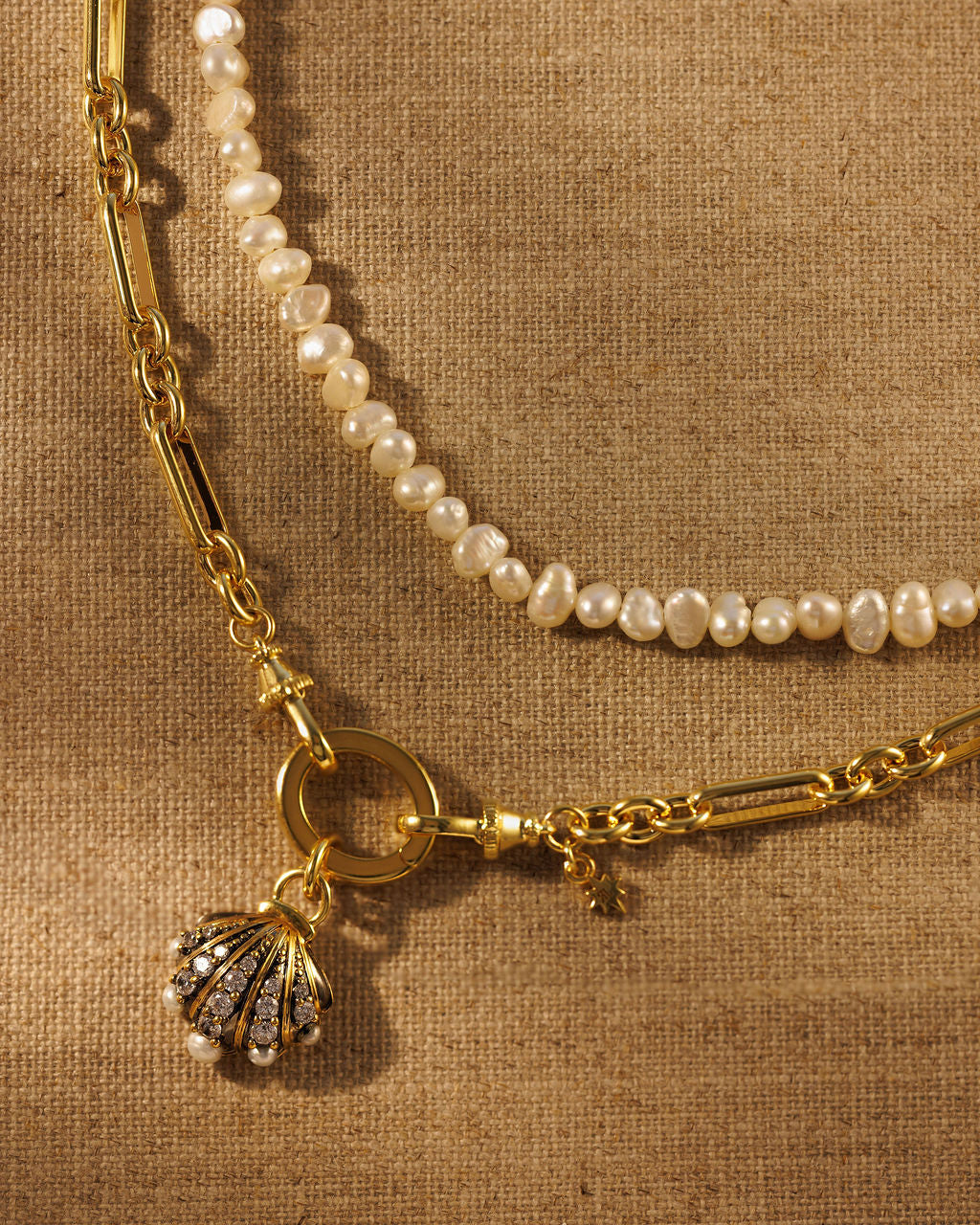 A close-up of a layered gold necklace set on a textured beige fabric background. The necklace features a gold chain with a shell-shaped pendant studded with small clear stones and white pearls draped alongside it.