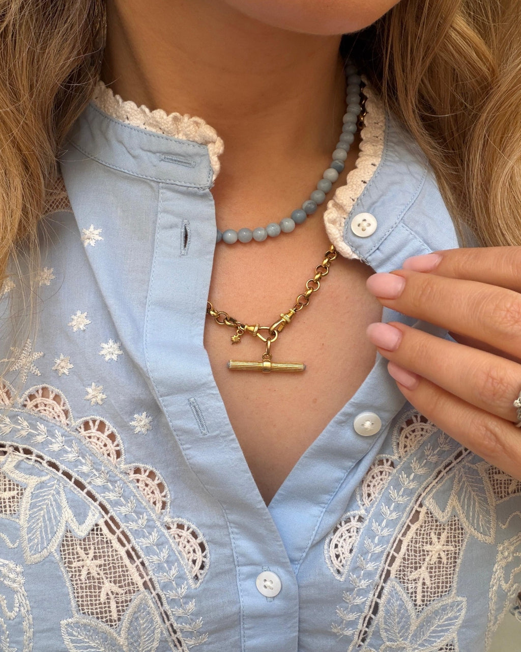 A close-up of a person wearing a gold t bar necklace layered over a light blue embroidered blouse with lace details. The person is also wearing a light blue beaded necklace.