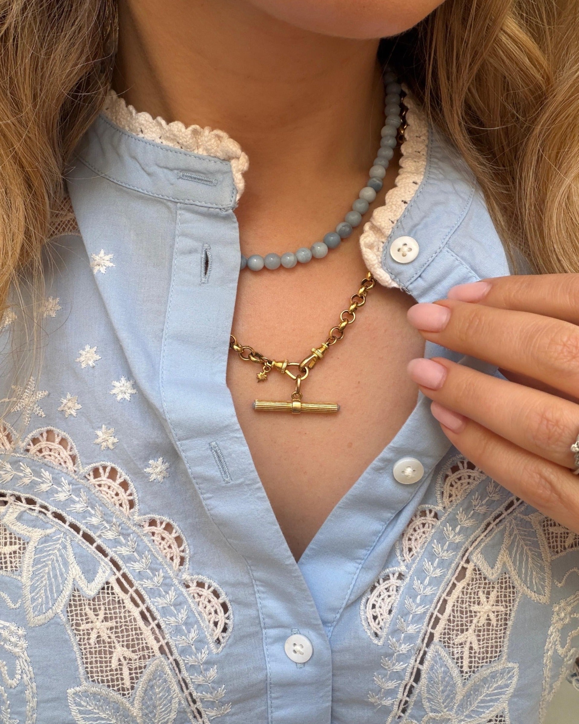 A close-up of a person wearing a gold t bar necklace layered over a light blue embroidered blouse with lace details. The person is also wearing a light blue beaded necklace.